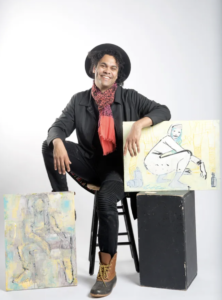 Artist Bryan Moss sits on a brown stool. He has brown skin and is wearing a black suit with a red scarf. His hand rests on some artwork.