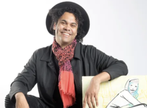 Artist Bryan Moss sits on a brown stool. He has brown skin and is wearing a black suit with a red scarf. His hand rests on some artwork.