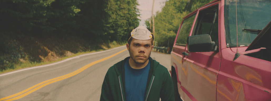 A man, wearing a K95 mask atop his head, stares grimly ahead. He wears a green hoodie and stands in the middle of a road next to a red car.