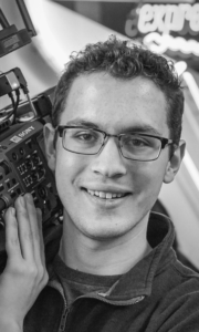 Black and white image of filmmaker Aaron Connor with camera on shoulder. He's wearing a zipped pullover and glasses.