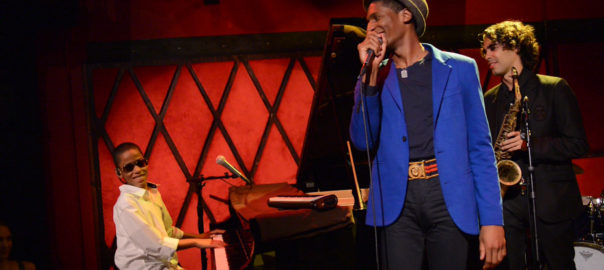 A man with a microphone stands in front of a boy playing a piano