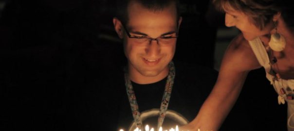 A man with glasses sits while a woman places a birthday cake with lit candles in front of him
