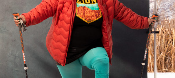 A woman stands in snow in a bright red jacket, holding ski poles. She has one foot, which is a prosthesis, on a box.
