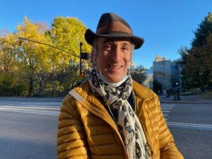 A man wearing a hat, yellow jacket and long scarf smiles at the camera.