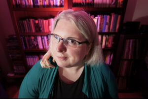 a blond woman with glasses smirking at the camera, with a prostethic hand placed on her sholder