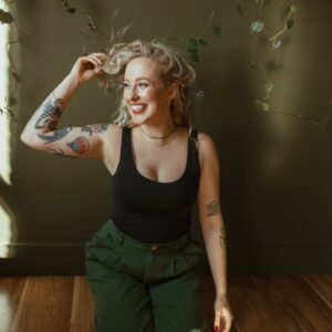 A young blong woman, leaning against a brown sofa, touching her hair, tattoos on her arm