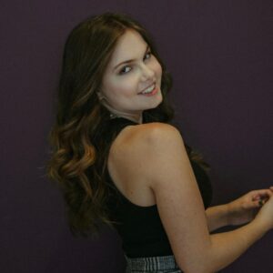 a young girl with long waivy brown hair, wearing a sleevless black top, looking sideways to the camera, smiling