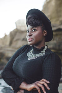 A black woman dressed in black, with a large silver neckless, wearing a black hat