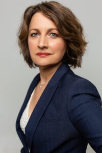 a woman wearing a blue blazer jacket, with light brown hair above the shoulders, looking sideways at the camera