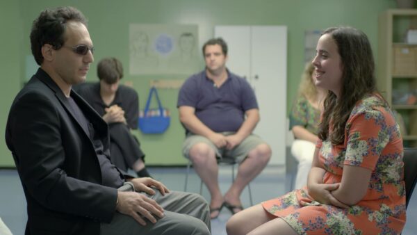 A girl in an orange floral dress sits across from a man in a suit with dark glasses. Other group members sit in the background.