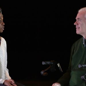 A silver haired white man sits in front of a black background, looking at a young black woman in white with a floral headwrap.