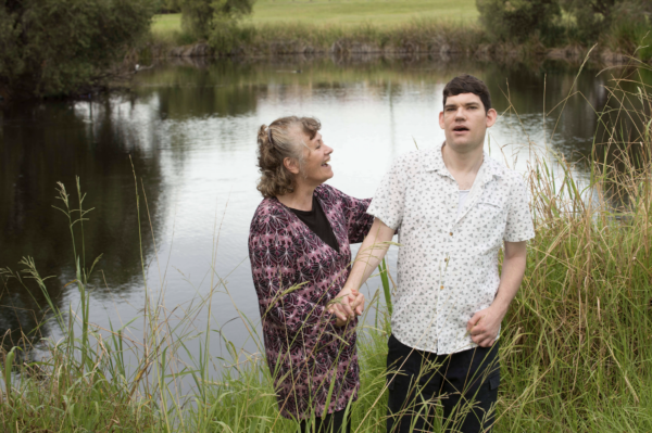 A young man in a white shirt stands next to an older woman in purple, who looks at him and touches his shoulder, smiling. The two stand in front of a pond with grass in the foreground of the shot.