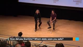 Two people sit on wooden stools on a stage facing an audience.