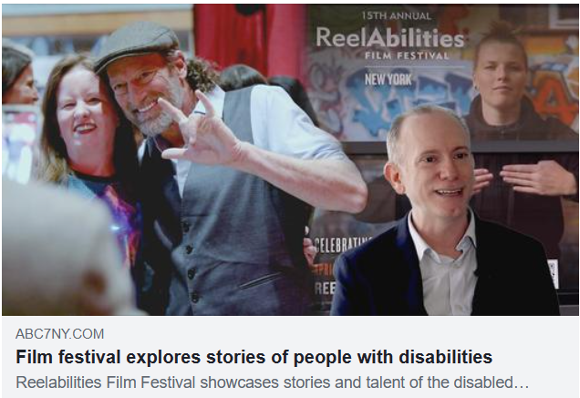 A man in a gray cap and vest with gray facial hair hugs a woman with light brown hair from the side. In the foreground, we see a man in a suit in front of a ReelAbilities poster.