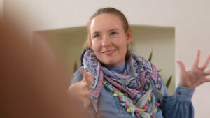 A young white girl with a dirty blonde ponytail sits with her hands up and fingers flexed as she smiles offscreen to the left. She wears a blue dress and a colorful scarf.