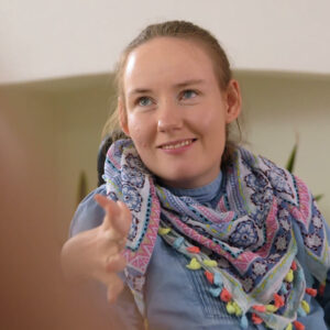 A young white girl with a dirty blonde ponytail sits with her hands up and fingers flexed as she smiles offscreen to the left. She wears a blue dress and a colorful scarf.