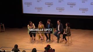 A panel of five people sit on wooden stools on a stage, holding microphones. Two men and three women participate.