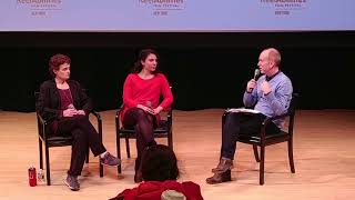 Two women sit on a well-lit stage with microphones. To their right, a man moderates.