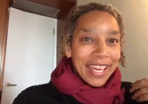 A brown skinned woman with light curly hair and a fuchsia scarf smiles at the camera.