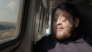 A bearded man with facial differences is sitting on the train looking out the window and smiling, while the sunlight hits his face.