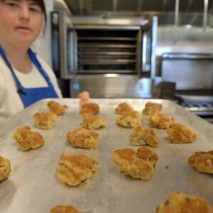 A young woman with Down syndrome wearing a blue apron holds a tray of freshly baked goods in a commercial kitchen. An open oven is in the background.