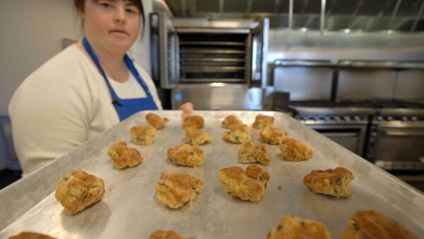 A young woman with Down syndrome wearing a blue apron holds a tray of freshly baked goods in a commercial kitchen. An open oven is in the background.