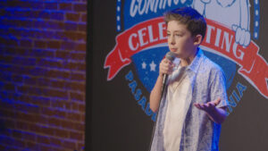 A young light-skinned person in a patterned shirt telling jokes on a comedy stage with a microphone.