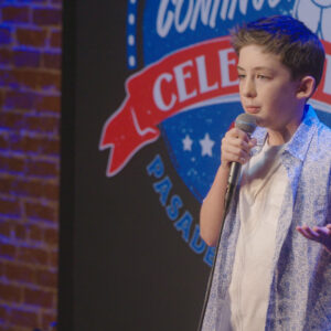 A young light-skinned person in a patterned shirt telling jokes on a comedy stage with a microphone.