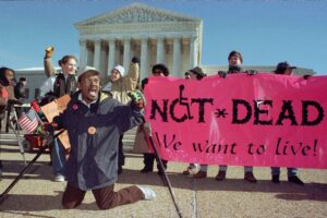 A group of disability rights activists protest in front of the Supreme Court, holding a pink banner that reads "NOT * DEAD - We want to live!". A man with a crutch kneels in front of them.