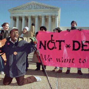 A group of disability rights activists protest in front of the Supreme Court, holding a pink banner that reads "NOT * DEAD - We want to live!". A man with a crutch kneels in front of them.