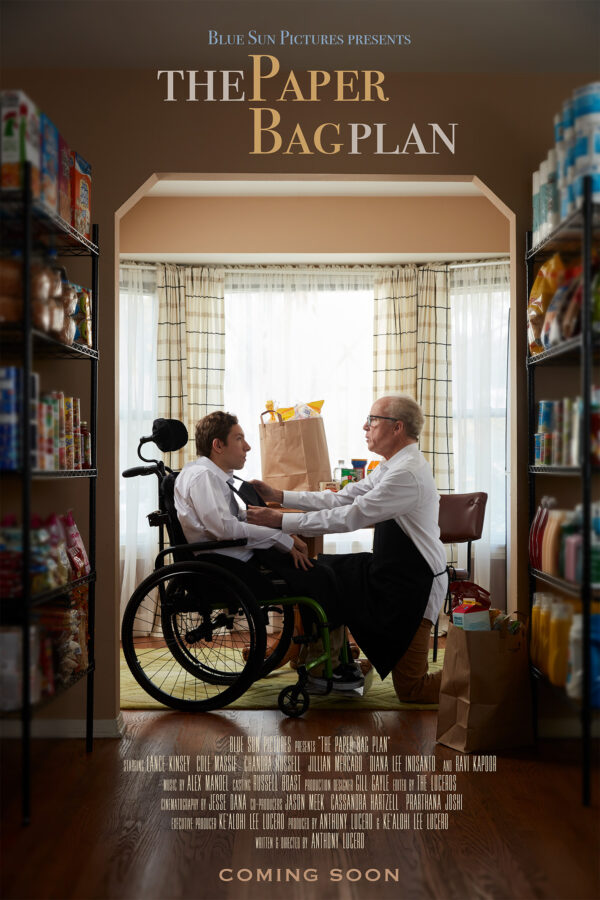 A movie poster for the film “The Paper Bag Plan” features a young man with dark hair in a wheelchair, wearing a white dress shirt. He is being assisted by an older man with glasses, who is dressed in a shirt and apron. They are surrounded by shelves filled with food and positioned in front of a window.