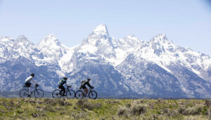 A wide shot from the movie "Blind AF" captures three cyclists riding across a grassy field with snow-capped mountains in the background. The sky is clear and blue.