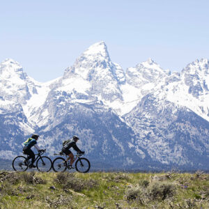 A wide shot from the movie "Blind AF" captures three cyclists riding across a grassy field with snow-capped mountains in the background. The sky is clear and blue.