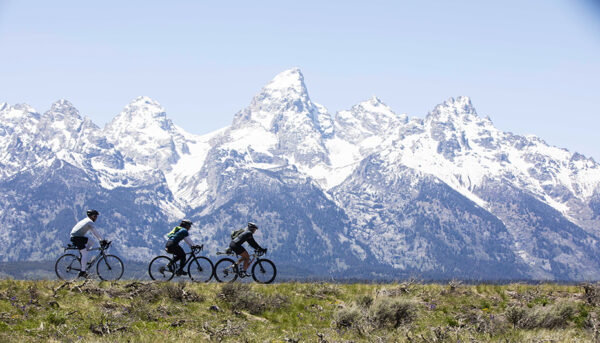 A wide shot from the movie "Blind AF" captures three cyclists riding across a grassy field with snow-capped mountains in the background. The sky is clear and blue.