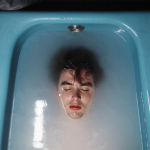 A person with closed eyes, dark hair, prominent dark eyebrows and red lips is partially submerged in water in a blue bathtub.