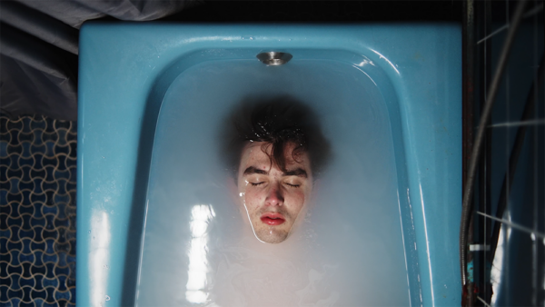 A person with closed eyes, dark hair, prominent dark eyebrows and red lips is partially submerged in water in a blue bathtub.