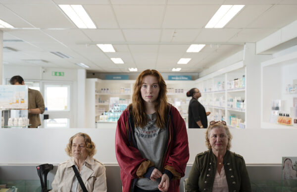 A young red-haired woman stands in a pharmacy surrounded by shelves of products, with other customers seated and standing in the background