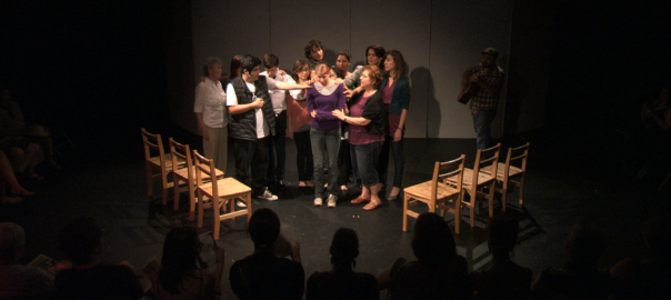 A group of people gather in a circle on a dark stage with wood chairs