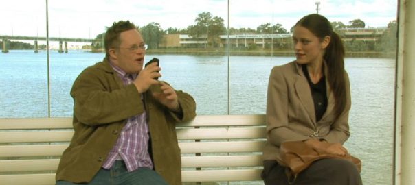 A man and a woman sit on a park bench. There is a body of water behind them