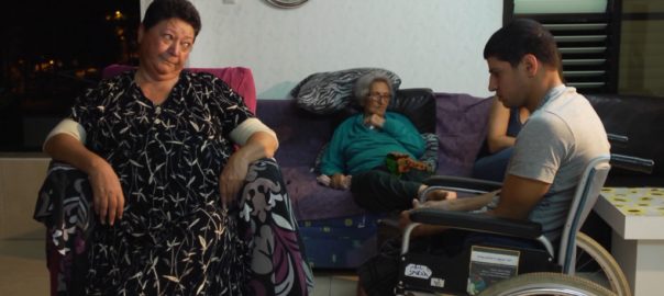 A woman sits on a couch. Two people in wheelchairs are in front of her