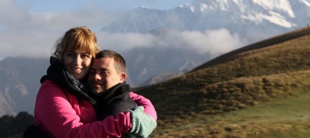 A man and woman hug in a green field with mountains behind them