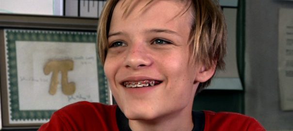 A boy smiles. a framed image of a Pi symbol is behind him