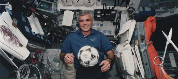 An astronaut in a rocket has a soccer ball floating in front of him