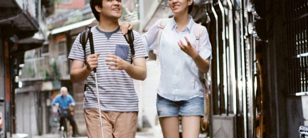 A man and woman walk together in a street. The man has a long support cane