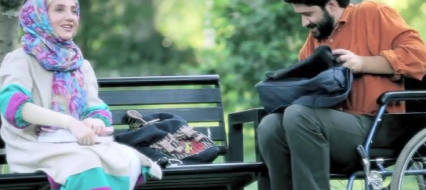 A woman sits on a park bench. A man in a wheelchair is next to her