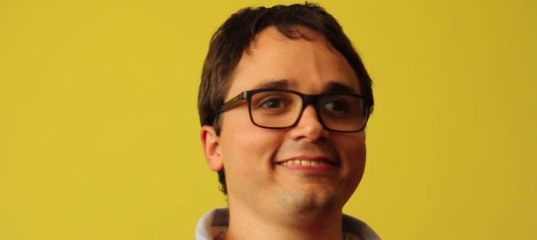 A man with glasses stands in front of a yellow background