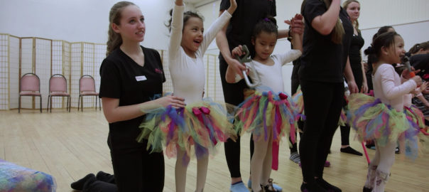A group of children in ballerina outfits stand with adults