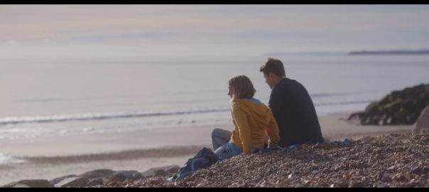 A man and woman sit on a hill above a beach