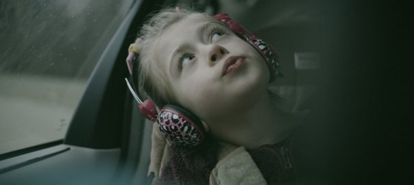 A young girl with headphones leans against a car window looking up