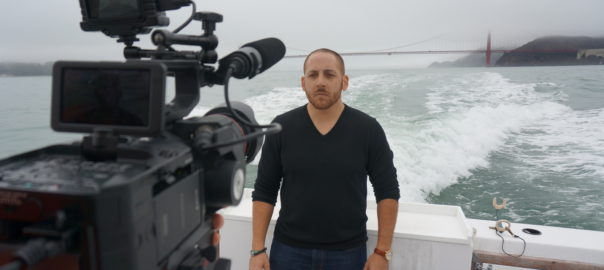 A man stands on the back of a moving boat. A camera is pointed towards him